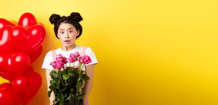 Happy Valentines day. Surprised asian girl receive bouquet of pink roses from lover, looking with awe at camera, standing near red heart balloons, yellow background.