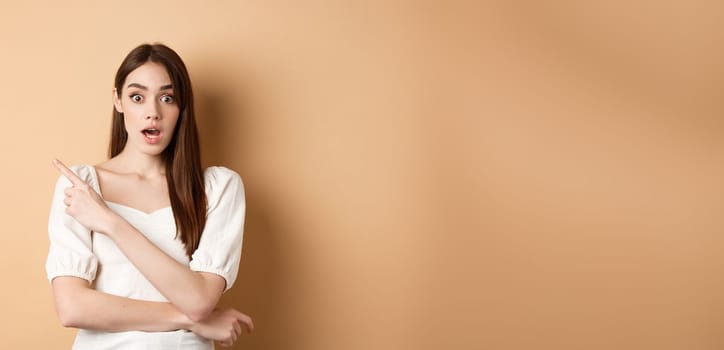 Surprised young girl say wow, pointing finger left at interesting banner, standing curious against beige background.