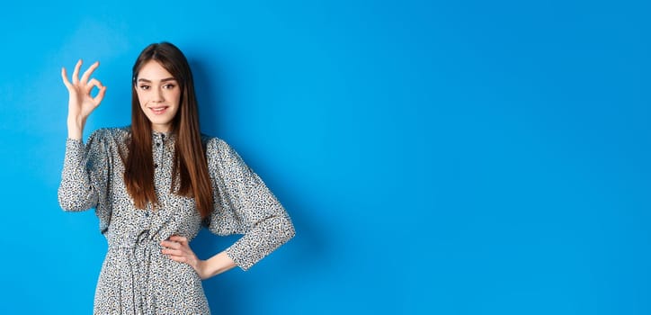 Confident young woman in dress showing okay sign and smiling, no problem gesture, assure everything good, standing on blue background.