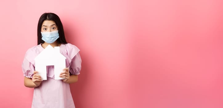 Real estate and pandemic concept. Excited asian girl in medical mask showing paper house cutout, standing against pink background.