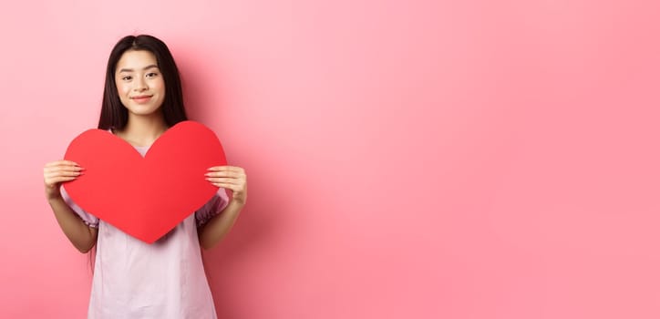 Valentines day concept. Cute teenage asian girl showing big red heart card, falling in love, going on romantic date in dress, smiling tender at camera, pink background.