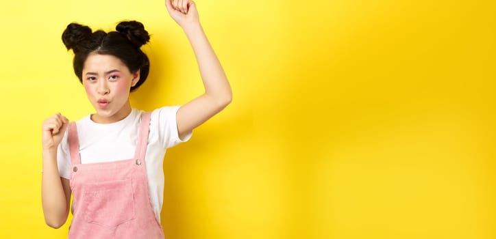 Excited asian woman getting motivation, raising hand up and chanting, celebrating victory, triumphing and standing on yellow background.