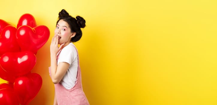 Valentines day and relationship concept. Coquettish and romantic girl laughing, covering mouth with hand, look silly at camera, standing near red heart balloons, yellow background.