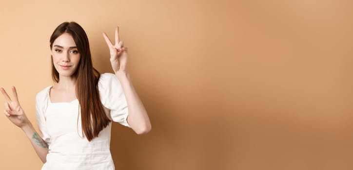 Cheerful attractive girl dancing and having fun, showing v-signs and looking positive at camera, standing on beige background.