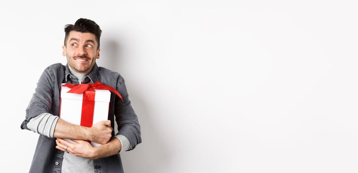 Happy boyfriend hugging gift from lover, looking aside at empty space and smiling excited, making romantic present on valentines day, standing on white background.