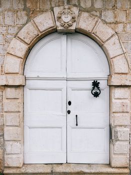 White arched wooden door in a stone wall with a carved knocker and a coat of arms above the entrance. High quality photo