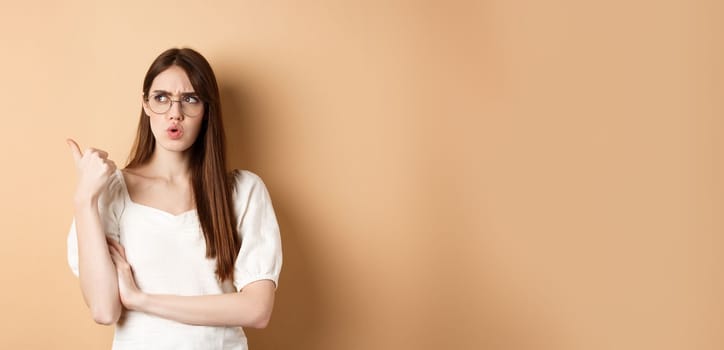 Confused and suspicious young woman in glasses frowning, pointing and looking aside with displeased face expression, standing on beige background.