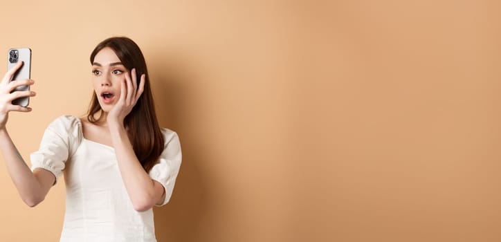 Fashion girl record smartphone blog, taking selfie on cellphone, standing on beige background.