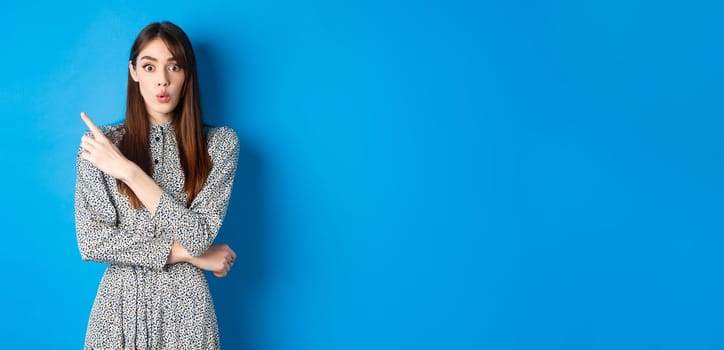 Excited young woman say wow, looking impressed and pointing left at logo, showing awesome news, standing cute dress on blue background.