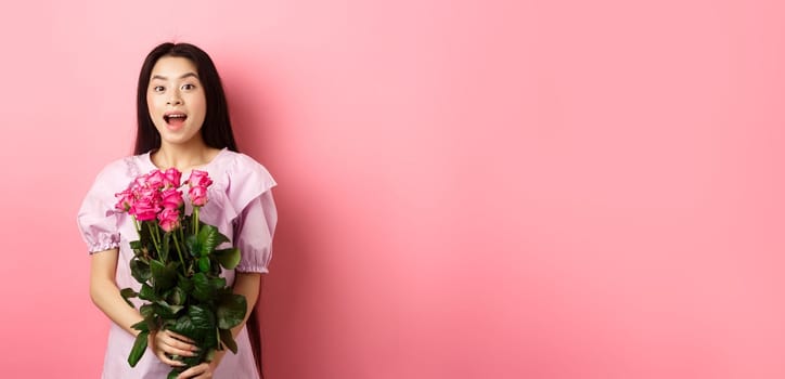 Korean teen girl in dress having romantic date on Valentines day, holding bouquet of roses and looking surprised at camera, receive gift on date from lover, pink background.