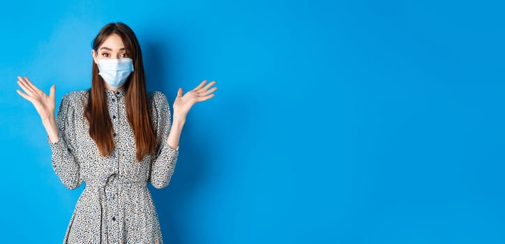 Healthy people and covid-19 pandemic concept. Surprised girl in medical mask raising hands up excited, hear good news, smiling with eyes, standing blue background.