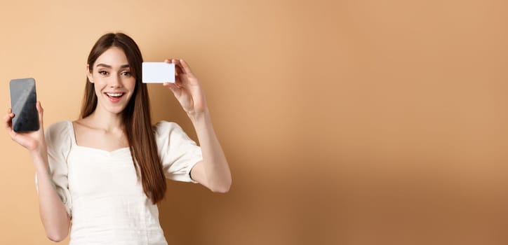 Cheerful young woman showing plastic credit card and empty mobile phone screen, smiling pleased at camera, standing on beige background.