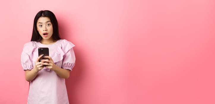 Online shopping. Surprised asian woman in cute dress, open mouth amazed after reading news on smartphone, standing on pink background.