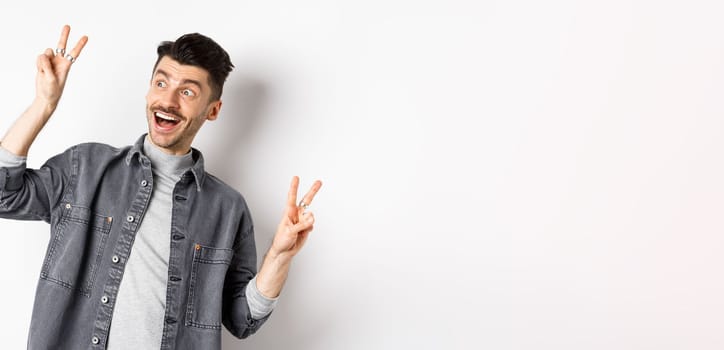 Funny young man showing v-signs and smiling at left side, looking at empty space with cheerful face expression, standing on white background.