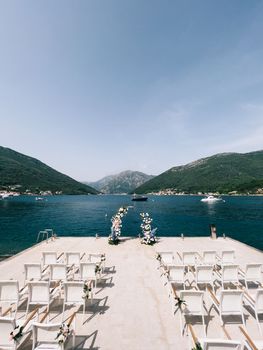 Rows of chairs decorated with flowers stand on the pier near the wedding semi-arch. High quality photo