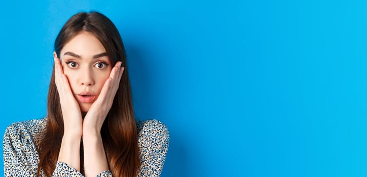Close-up portrait of surprised cute woman holding hands on cheeks and gasping, stare at camera shocked, standing against blue background.