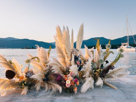Wedding arch made of dried flowers stands on a pier by the sea against the backdrop of mountains. High quality photo