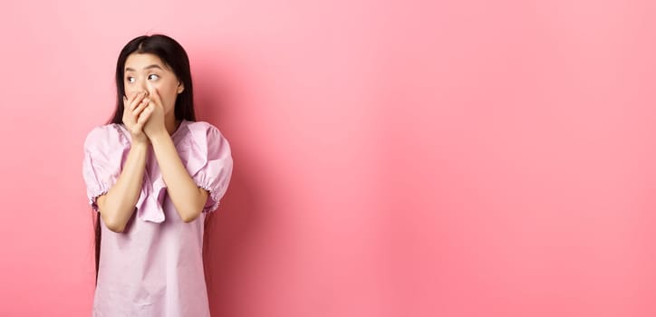 Shocked and startled asian woman looking aside at logo, covering mouth with hands speechless, standing in dress on pink background.