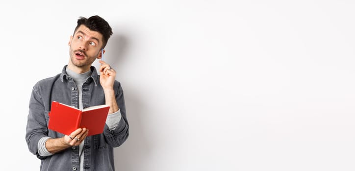Image of pensive young man writing down ideas in planner, looking thoughtful at logo and scratching ear with pen, holding journal in hands, white background.