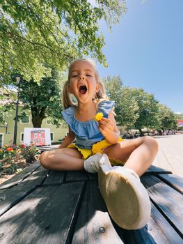 Little girl in blue and yellow clothes with a flower in her hand sits on a wooden table in the park. High quality photo