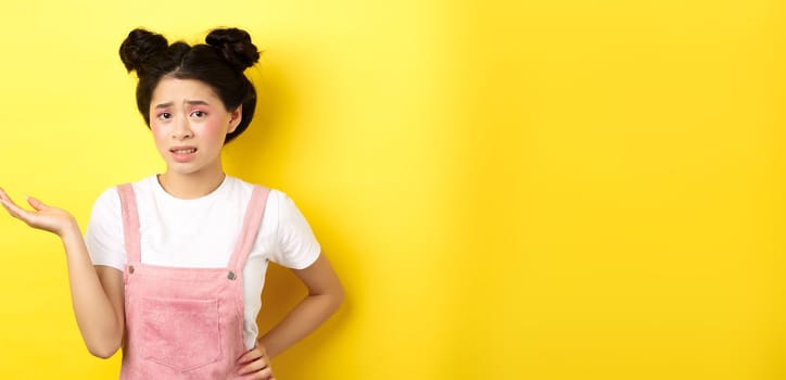 Confused teen asian girl with bright makeup and summer clothes, raising hand up and frowning puzzled, cant understand, yellow background.