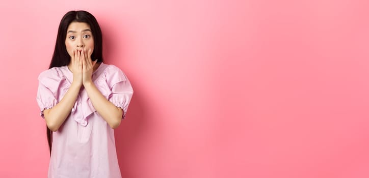 Shocked asian girl with long dark hair, gasping and covering mouth with hands, look at bad news, terrible accident, standing on pink background.