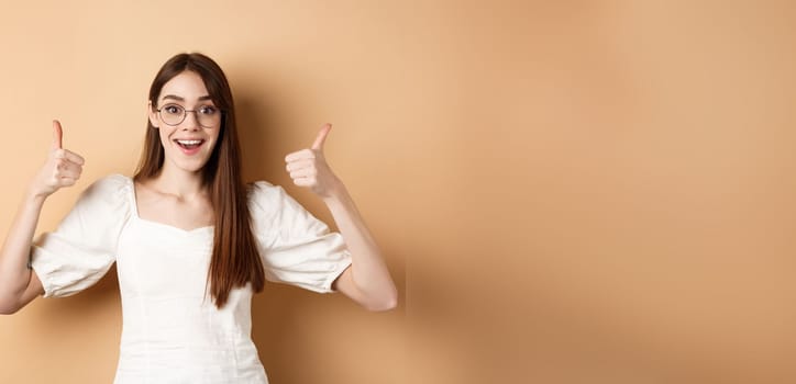 Excited young woman in glasses showing thumbs up and smiling, praising good optic store, approve and like eyewear, standing on beige background.