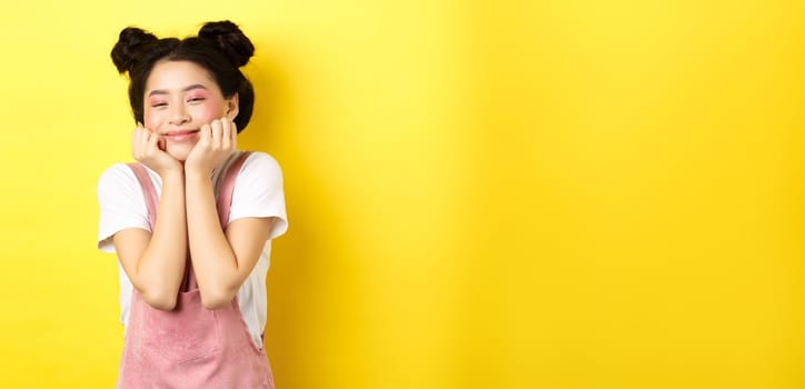 Cute happy asian woman with bright makeup and dungarees, leaning satisfied on hands and smiling, standing on yellow background.