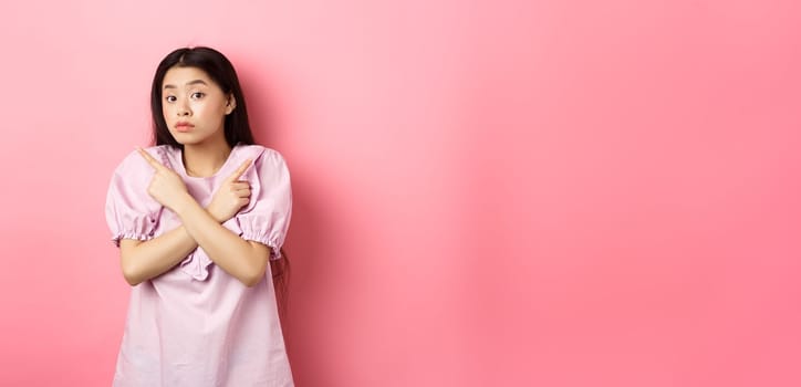 Clueless cute asian girl pointing sideways and look confused, cant make decision, standing hesitant against pink background.