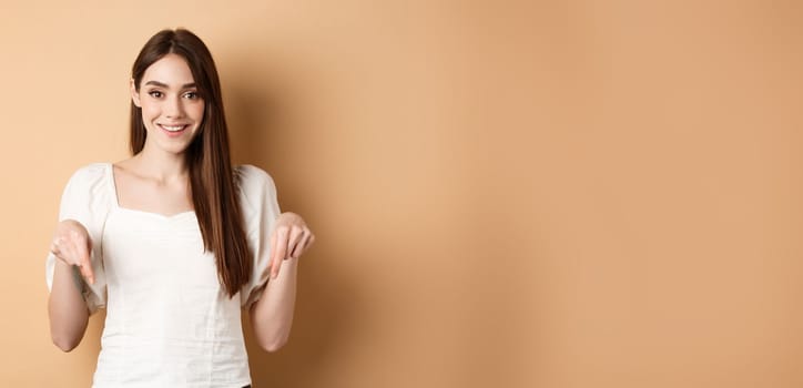 Cheerful young girl with lovely smile, pointing fingers down, making announcement or inviting click link, showing logo, beige background.