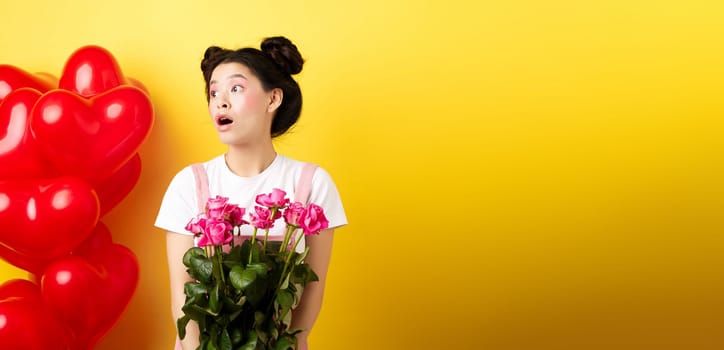 Happy Valentines day. Surprised asian girl looking left with silly face, holding cute pink flowers, receive romantic bouquet, standing on yellow background near red hearts balloons.