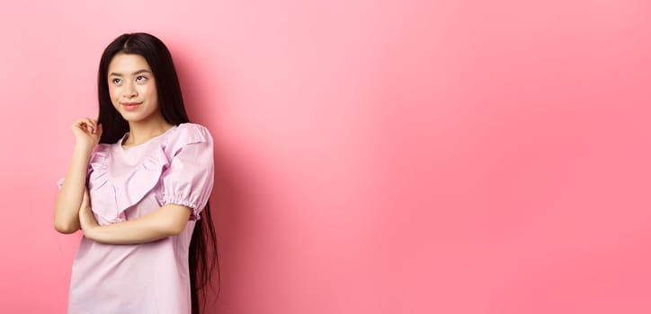 Beautiful asian woman smiling and looking aside, standing in dress on pink romantic background.