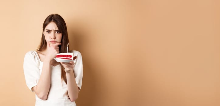 Thoughtful birthday girl frowning and thinking of wish, holding cake with candle, standing on beige background.