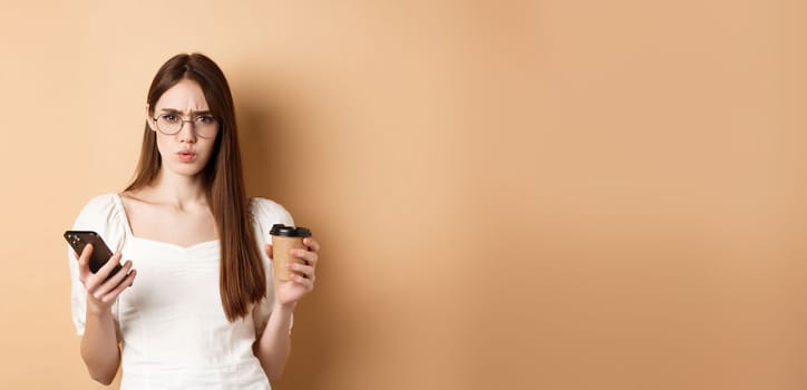 Confused girl in glasses frowning at camera after reading mobile phone, holding coffee cup, standing displeased on beige background.
