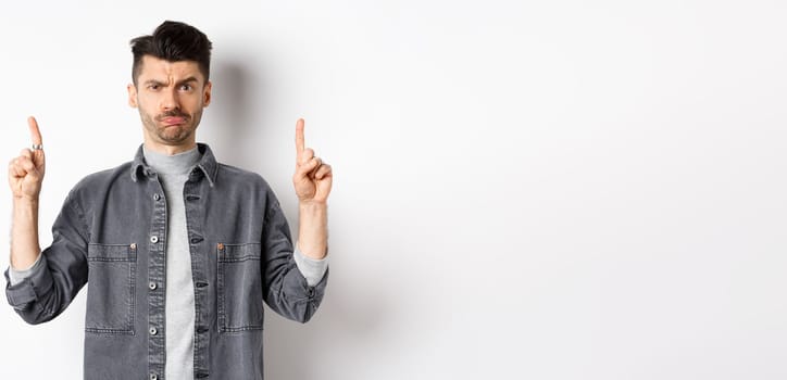 Confused funny guy with moustache pointing fingers up at something strange, frowning and pouting puzzled, standing on white background.