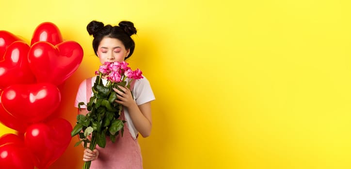 Happy Valentines day. Tender asian girl smelling flower from boyfriend. Girlfriend holding beautiful pink roses, standing near romantic hearts balloons, yellow background.