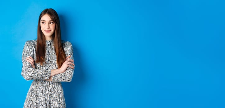 Smiling woman in romantic dress cross arms on chest and looking left at logo, standing against blue background.