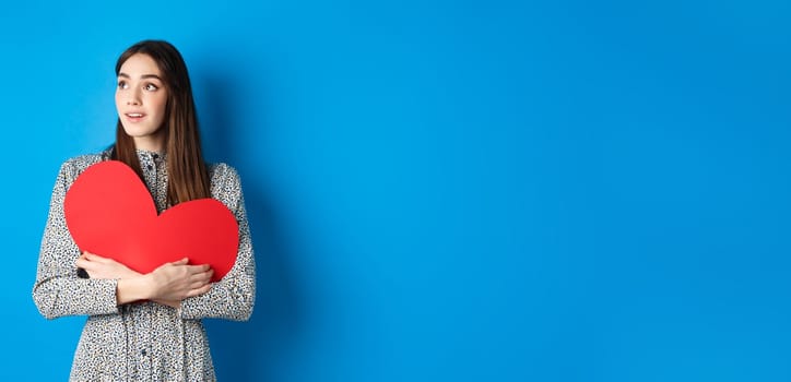 Valentines day. Hopeful girl dreaming off soulmate, looking aside at empty space with excitement, hugging big red heart cutout, standing on blue background.