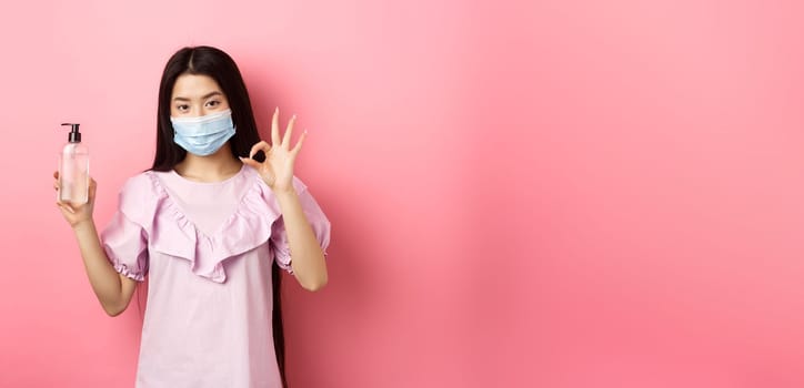 Healthy people and covid-19 pandemic concept. Cheerful asian woman in medical mask recommend hand sanitizer, showing okay sign, good antiseptic bottle, white background.