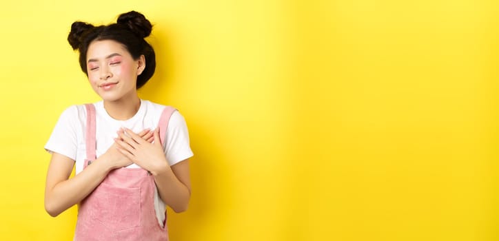 Romantic asian girl with makeup, wearing summer clothes, holding hands on heart and smiling tenderly, remember tender memory, standing on yellow background.