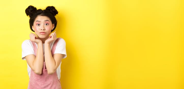 Cute teenage asian girl with beauty makeup, listen with interest, lean on hands and looking excited at camera, standing on yellow background.