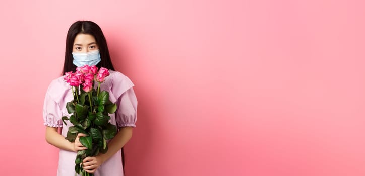 Young asian girl in medical mask holding flowers on Valentines day, receive bouquet of roses from lover, standing on pink background. Social distancing and covid-19 concept.
