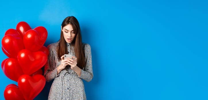 Valentines day. Portrait of young woman standing near red romantic balloons, looking surprised at smartphone screen, blue background.