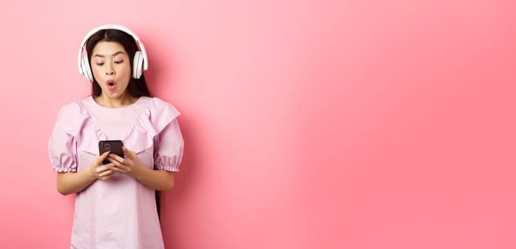 Excited asian girl looking at smartphone screen amused, say wow, listening music in wireless headphones, standing against pink background.