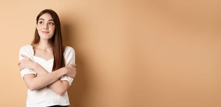 Romantic young girl dreaming, hugging herself and smiling at upper left corner, imaging something lovely, standing on beige background.