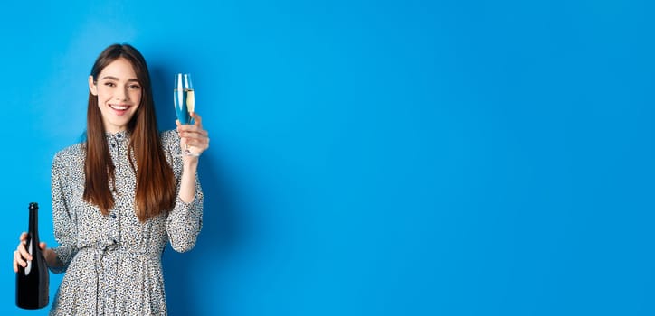 Celebration. Happy young woman in dress raising glass of champagne, holding bottle and smiling, having fun at party, standing on blue background.