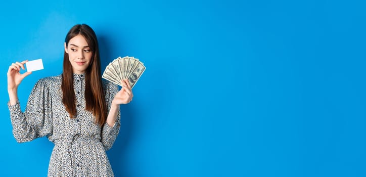Shopping. Beauty girl in dress looking aside and smiling pensive, showing money and plastic credit card, standing on blue background.