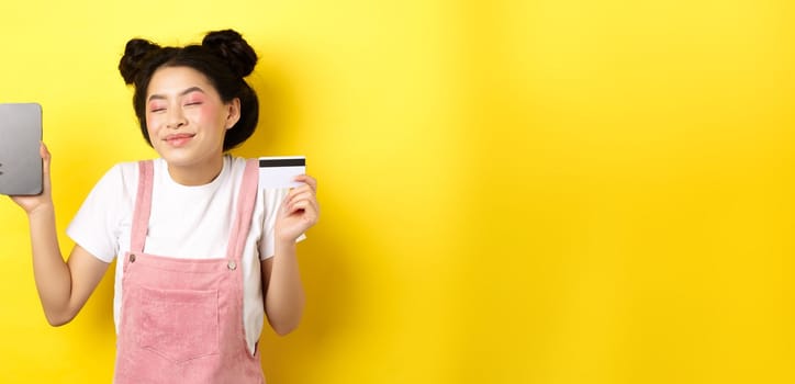 Online shopping concept. Happy asian girl showing empty smartphone screen and credit card, paying contactless, standing on yellow background.