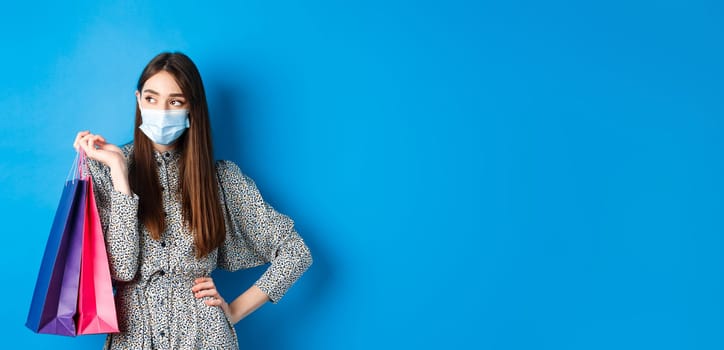 Covid-19, pandemic and lifestyle concept. Girl wearing medical mask on shopping in malls, holding paper bags and look aside at empty space, standing on blue background.