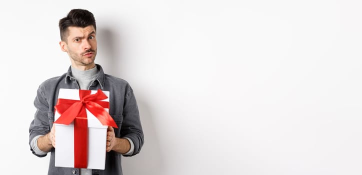 Surprised young man look with disbelief and hold surprise gift, raising eyebrow doubtful, being suspicious, standing on white background. Valentines day concept.